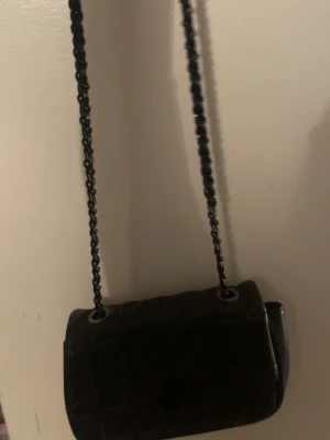 Svart quiltad axelväska med kedja - Säljer en svart quiltad axelväska med lång kedjerem från Even&Odd. Väskan har en klassisk rektangulär form och stängs med ett runt lock. Perfekt för dig som gillar stilrena detaljer och vill ha en väska som sticker ut lite extra.