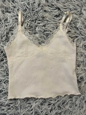 Vit ribbad linnetopp med spets - Säljer en söt vit linnetopp med tunna axelband och ribbad struktur. Toppen har fina spetsdetaljer längs urringningen och under bysten, vilket ger en romantisk känsla. Perfekt att styla med jeans eller kjol för en fräsch look.