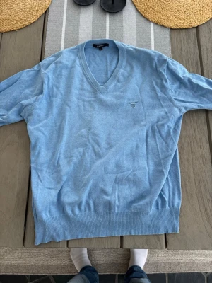 Ljusblå v-ringad tröja från Gant L - Säljer en ljusblå v-ringad tröja från Gant med diskret logga broderad på bröstet. Tröjan har långa ärmar, ribbade muddar och är tillverkad i mjuk bomull. Perfekt för dig som gillar klassisk och stilren look. Storlek L