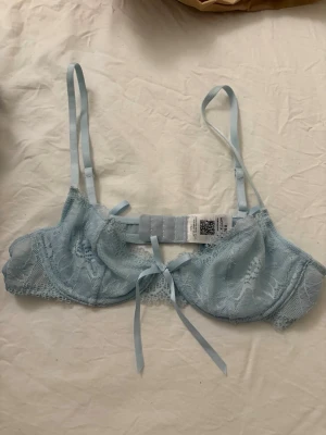 Ljusblå spets bh med rosetter Nelly - Säljer en ljusblå, tunn och jättesöt spets bh från Nelly med flera dekorativa rosetter framtill och baktill. Bh:n har justerbara axelband och fina spetsdetaljer som ger en feminin look. Perfekt för att addera något extra till din outfit. Aldrig använd endast testad, säljer då den var för liten för mig!