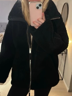 Svart teddyjacka med dragkedja - Mysig svart teddyjacka med oversized passform och stora fickor framtill. Jackan har en hög krage och grov silverfärgad dragkedja hela vägen. Perfekt för kalla dagar när du vill vara både varm och snygg. Materialet är fluffigt och mjukt, vilket ger en riktigt skön känsla.