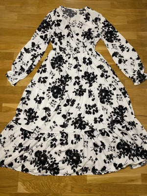 Vit långklänning med svart blommönster - Säljer en vit långklänning med svart blommigt mönster. Klänningen har omlott-v-ringning, långa ärmar med resår vid handlederna och markerad midja. Tyget är mjukt och faller snyggt, troligtvis i polyester. Perfekt för dig som gillar romantisk och stilren look.