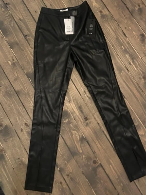 Svarta PU-byxor från NA-KD, strl 36 - Snygga svarta PU-byxor från NA-KD i storlek 36. Byxorna har raka ben med slits framtill vid benslut. Helt nya med prislapp kvar. 
