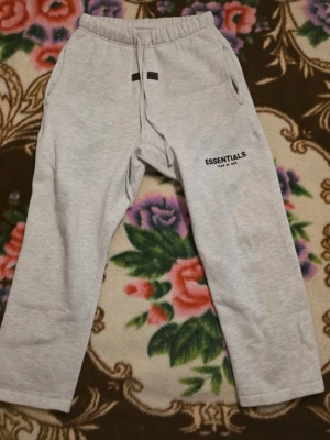Grå Essentials mjukisbyxor Fear of God - Snygga ljusgrå mjukisbyxor från Essentials Fear of God med svart logga på benet. Byxorna har dragsko i midjan, två sidofickor och en relaxed passform. Perfekta för chill dagar eller streetwear-stil.