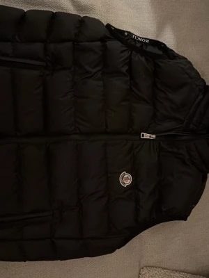 Svart dunväst från Moncler - Snygg svart dunväst från Moncler med klassisk quiltad design och dragkedja framtill. Västen har Monclers ikoniska logga på bröstet och en diskret bröstficka med dragkedja. Perfekt för lager-på-lager och ger en clean streetwear-look.