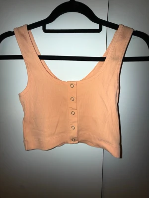 Ljusrosa ribbad linnetopp med knappar - Snygg ljusrosa ribbad linnetopp med knappar framtill. Croppad modell med breda axelband och rundad halsringning. Perfekt till sommaren och enkel att matcha med jeans eller kjol.