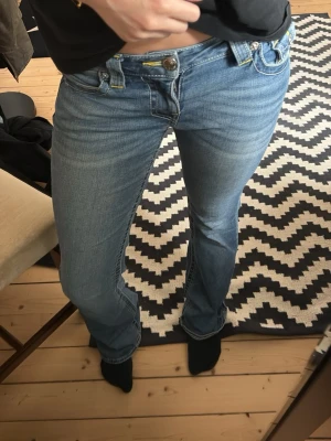 Blå bootcut jeans med fickdetaljer - Snygga blå bootcut jeans från True religion med dekorativa fickor bak och kontrastsömmar. Jeansen har låg midja och klassisk femficksdesign. Perfekta för dig som gillar en avslappnad men trendig look. Köpta för 1500kr säljer för 600!!!