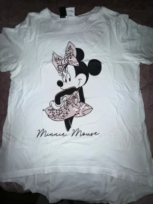 Vit Minnie Mouse t-shirt - Vit t-shirt med tryck av Minnie Mouse i svart och rosa. Motivet har glittriga detaljer på rosett och klänning. T-shirten har rund hals och korta ärmar, perfekt för dig som gillar Disney och vill ha en söt och lekfull stil.