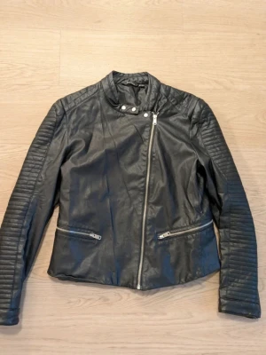 Svart bikerinspirerad skinnimitation  - Snygg svart skinnimitationjacka med bikerstil och quiltade detaljer på ärmarna. Jackan har sned dragkedja, två fickor med dragkedja och ståkrage med tryckknappar. Perfekt för en cool och stilren look.