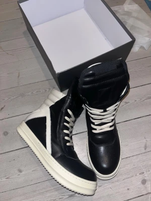 Svarta Rick Owens geobaskets med vita detaljer - Säljer ett par svarta och vita high top sneakers från Rick Owens. Skorna har en chunky vit sula, vita snören och detaljer i vitt skinn. Klassisk rund tå och högt skaft som ger en edgy look. Perfekta för dig som gillar streetwear och vill sticka ut.
