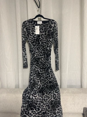 Leopardmönstrad långklänning Sisters Point - Snygg långklänning från Sisters Point i svart och grått leopardmönster. Klänningen har lång ärm och figurnära passform med rynkade detaljer längs sidorna. Tillverkad i mjukt stretchigt material som ger en bekväm känsla.