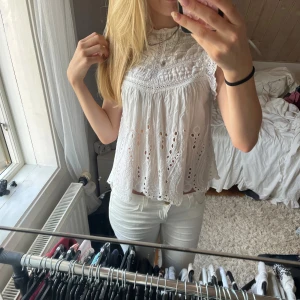 Vit broderad blus med spetsdetaljer - Supersöt vit blus med broderade detaljer och spets längs kanterna. Blusen är ärmlös och har en luftig, lätt A-linjeform med fina hålmönster och hög hals. Perfekt för en fräsch och somrig look.