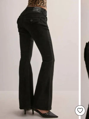 Svarta bootcut jeans från True Religion - Snygga svarta jeans från True Religion med bootcut passform och ikoniska bakfickor med lock och knapp. Jeansen har hög midja och klassisk jeanslook med tydliga sömmar och logopatch bak. Perfekta för dig som vill ha en cool och tidlös stil.
