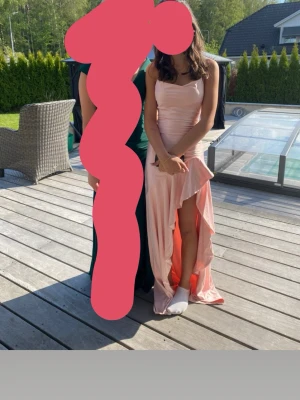 Ljusrosa balklänning - Säljer en ljusrosa långklänning med asymmetrisk volangkant och slits framtill. Klänningen är ärmlös och har en figurnära passform som framhäver siluetten. Perfekt för sommarens speciella tillfällen.