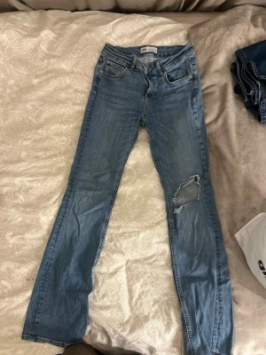 Blå raka jeans med slitning från Zara - Säljer ett par ljusblå jeans från Zara med rak passform och en snygg slitning på höger knä. Klassisk femficksmodell och normal midja. Perfekta till vardags för en avslappnad look.