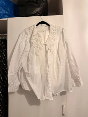 Vit blus med spetskrage - Säljer en vit blus med långärmad passform och en stor dekorativ spetskrage. Blusen har knäppning framtill och är gjord i ett lätt tyg som ger en luftig känsla. Perfekt för dig som gillar romantiska detaljer och vill sticka ut lite extra.
