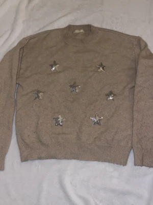 Beige stickad tröja med paljettstjärnor - Säljer en beige stickad tröja med rund hals och långa ärmar. Tröjan är dekorerad med flera silvriga paljettstjärnor på framsidan som ger en unik och trendig look.