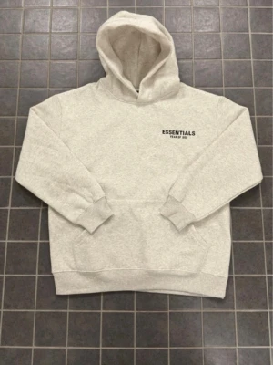 Beige Essentials Fear of God hoodie - Snygg beige hoodie från Essentials Fear of God med svart logga på bröstet och stor text på ryggen. Hoodien har en stor huva, ribbade muddar och en klassisk känguruficka framtill. Perfekt för en avslappnad och trendig streetwear-look.