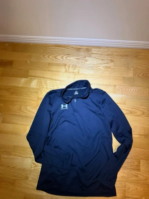 Svart långärmad träningströja UA - Svart långärmad träningströja från Under Armour med halv dragkedja och hög krage. Tröjan har en diskret logga på bröstet och är tillverkad i ett stretchigt funktionsmaterial som andas. Perfekt för träning eller löpning.