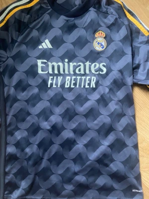 Real Madrid bortatröja Adidas - Äkta Mörkblå Real Madrid bortatröja från Adidas med gulddetaljer på axlarna och ett unikt mönster över hela tröjan. Broderad klubbmärke på bröstet och 'Emirates Fly Better' tryckt framtill. Tillverkad i lätt och ventilerande material. Är i väldigt bra skick och har inte använts alls nästan, säljer den för jag inte gillar Real Madrid. (Bellingham på ryggen.) Storlek 176 och är anpassade för 15-16 åringar. 