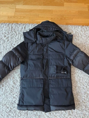 Svart dunjacka från C.P. Company - Snygg svart dunjacka från C.P. Company med flera stora fickor framtill och avtagbar huva. Jackan har lång ärm, dragkedja och tryckknappar. Perfekt för kalla dagar med sin pufferstil och moderna look. Köpt i Milano därav udda modell. Kontoutdrag/kvitto finns som gärna visas vid affär. Väldigt bra pris med tanke på nypris.