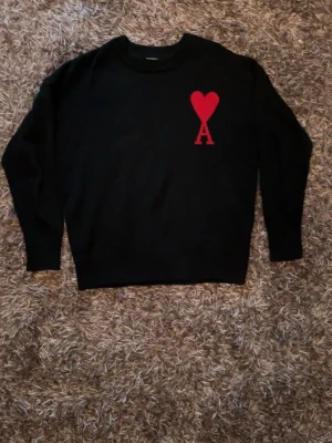 Svart stickad tröja från Ami Paris - Svart stickad tröja från Ami Paris med röd hjärtlogga och bokstaven A på bröstet. Tröjan har rund halsringning, långa ärmar och ribbade muddar vid ärmslut och nederkant. Perfekt för en stilren och trendig look.