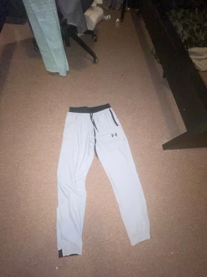 Grå mjukisbyxor från Under Armour - Snygga grå mjukisbyxor från Under Armour med svart midjeresår och snörning. Byxorna har en enkel design med logga på vänster ben och smal passform. Perfekta för chill eller träning.