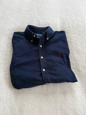 Mörkblå skjorta från Ralph Lauren - Klassisk mörkblå skjorta från Ralph Lauren med button-down krage och ikonisk röd broderad logga på bröstet. Tillverkad i mjuk bomull och har vita knappar framtill samt vid ärmsluten. Perfekt för en clean och stilren look.
