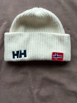 Vit ribbstickad mössa Helly Hansen - Vit ribbstickad mössa från Helly Hansen med uppvikt kant. Broderad HH-logga i svart och norsk flagga framtill. Klassisk och enkel design som passar till det mesta. Perfekt för kalla dagar. Oanvänd 
