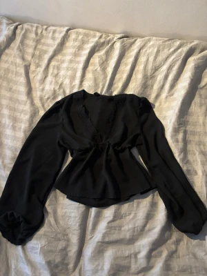 Svart omlottopp med spetsdetaljer - Snygg svart omlottopp med långa ärmar och v-ringning dekorerad med spets. Toppen har en lätt croppad passform och är tillverkad i ett tunt, mjukt material som ger ett elegant fall. Perfekt för dig som gillar stilrena och trendiga plagg.
