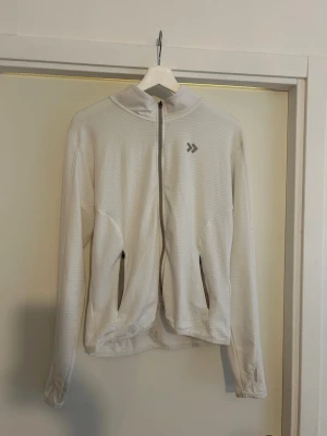 Vit ziptröja med hög krage - Vit ziptröja med hög krage och två fickor med dragkedja. Tröjan har ett diskret mönster i tyget och tumhål vid ärmsluten. Perfekt för lager-på-lager och en sportig look.