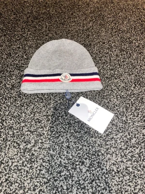 Grå Moncler mössa med ränder - Snygg grå ribbstickad mössa från Moncler med klassiska röda, vita och marinblå ränder på uppviket. Mössan har Moncler-logga framtill och är tillverkad i mjukt material som känns lyxigt. Perfekt accessoar för dig som vill ha en stilren och trendig look.