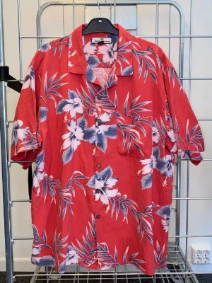 Röd blommig hawaiiskjorta - Snygg röd hawaiiskjorta med stora vita och blå blommor och gröna blad. Skjortan har korta ärmar, klassisk krage och knappar framtill. Perfekt för sommarens alla äventyr och ger en avslappnad vibe. 100% silke.