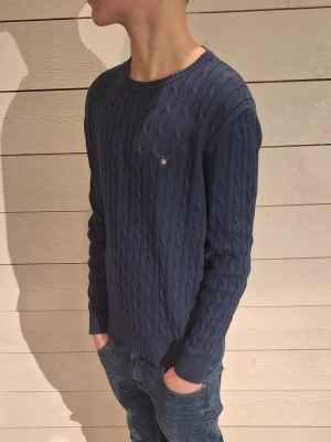 Marinblå kabelstickad tröja Gant - Säljer en marinblå kabelstickad tröja från Gant med rund hals och diskret logga på bröstet. Tröjan har lång ärm och ribbade muddar vid ärmslut och nederkant. Perfekt till jeans för en clean och klassisk look. Modelen är 162cm!