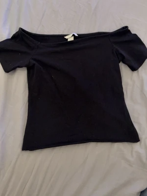 Svart offshoulder topp - En svart offshoulder topp med korta ärmar och figurnära passform. Enkel och stilren design som passar perfekt till jeans eller kjol. Mjuk och stretchig bomullskvalitet för en bekväm känsla.klippt på botten men ingen synlig skillnad från H&M