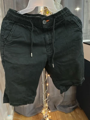 Svarta shorts från FSBN med snörning - Svarta shorts från FSBN med elastisk midja och snörning samt knapp och dragkedja. Modellen har sidofickor och är tillverkad i ett mjukt bomullsmaterial. Perfekta för en avslappnad och stilren look.