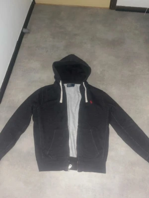 Mörkgrå hoodie från Polo Ralph Lauren - Snygg mörkgrå hoodie från Polo Ralph Lauren med dragkedja, vit insida och vita snören i huvan. Klassisk röd logga på bröstet och två fickor framtill. Perfekt för en chill och avslappnad stil.