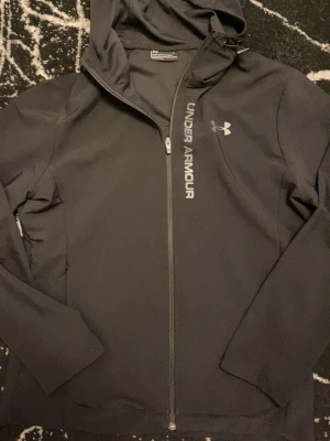 Svart vindjacka från Under Armour - Svart vindjacka från Under Armour med huva och hel dragkedja framtill. Jackan har logga och text på bröstet, samt en sportig och stilren design. Perfekt för blåsiga dagar och träning utomhus.