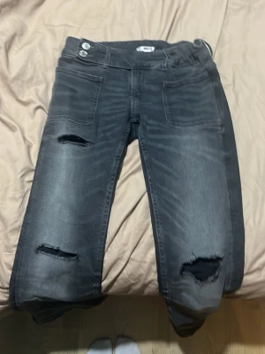 Svarta ripped jeans från Gina Tricot - Säljer ett par svarta jeans från Gina Tricot med slitningar på båda benen och stora framfickor. Jeansen har två knappar i midjan och en cool, lite tvättad look. Perfekta för dig som gillar en avslappnad och trendig stil.