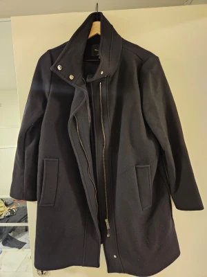 Svart ullkappa med hög krage - Stilren svart kappa från Max Mara i ull med hög krage och dold dragkedja framtill. Kappan har långa ärmar, två fickor och en rak passform. Perfekt för kyliga dagar när du vill vara både varm och snygg.