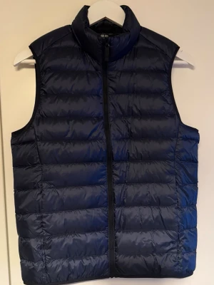 Mörkblå dunväst från Uniqlo XS(Ny utan prislapp) - Snygg mörkblå dunväst från Uniqlo i modellen Ultra Light Down. Västen har en hög krage, dragkedja framtill och är tillverkad i lätt, vattenavvisande material. Perfekt för lager på lager och enkel att bära med sig tack vare sin smidiga design.(Använd 1 gång)