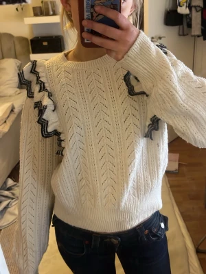 Vit stickad tröja med volang från Lindex - Säljer en vit stickad tröja sopm har snygga volangdetaljer med svarta och grå ränder på axlarna. Tröjan har ett hålmönster i zigzag och ribbade muddar vid ärmslut och nederkant. Perfekt för dig som vill ha något både gulligt och trendigt.