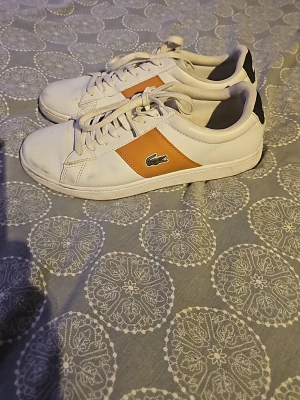 Vita sneakers från Lacoste - Snygga vita sneakers från Lacoste med en orange panel på sidan och klassisk krokodillogga. Skorna har snörning, rund tå och svarta detaljer vid hälen. Perfekta för dig som gillar stilrena och sportiga skor med en twist.