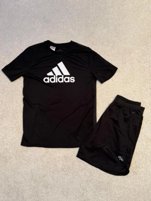 Svart Adidas/H&M träningsset - Säljer ett svart träningsset med en Adidas t-shirt i funktionsmaterial och stort vitt Adidas-tryck på bröstet. Samt matchande svarta H&M Move shorts i mjukt funktionsmaterial, med dragkedjefickor. Båda plaggen har en sportig och bekväm passform. Storlek, T-shirt:164. Shorts:XS. Båda i mycket fint använt skick.