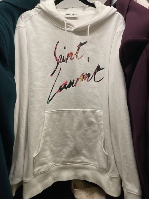 Vit hoodie från Saint Laurent - Säljer en vit hoodie från Saint Laurent Paris med färgglad logga framtill i graffiti-stil. Hoodien har huva med dragsko och en stor magficka. Tillverkad i mjukt bomullsmaterial som känns riktigt skönt mot huden. Perfekt för dig som vill ha en clean men ändå unik look.