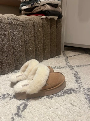 Bruna UGG tofflor med fårull - Mysiga bruna tofflor från UGG med fluffig vit fårull på insidan och kanten. Tofflorna har en platt sula och är perfekta för att hålla fötterna varma hemma. Klassisk UGG-logga på innersulan och mjukt mockamaterial på utsidan.