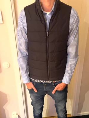 Svart dunväst från Bruun & Stengade - Snygg svart dunväst från Bruun & Stengade med hög krage och dragkedja framtill. Västen har en stilren, quiltad design och logotyp på ryggen. Perfekt att slänga över en skjorta eller hoodie för en clean look.