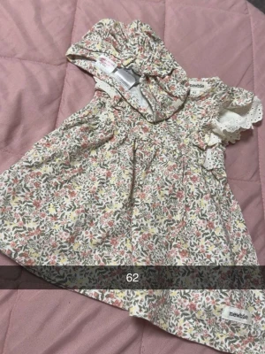 Blommig klänning från Newbie - Supersöt blommig klänning från Newbie med volangdetaljer och korta ärmar i broderad spets. Klänningen har en ljus bas med små blommor i rosa, grön och gul. Matchande mössa ingår. Perfekt för vår och sommar!