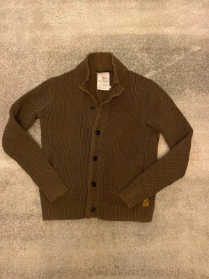 Mörkgrön cardigan/kofta från Dstrezzed - Säljer en mörkgrön stickad kofta från Dstrezzed med grov struktur, ståkrage och knäppning med svarta knappar. Två fickor framtill och ribbade muddar vid ärmslut och nederkant. Koftan är i L men passar M. Perfekt för alla årstider. Vid funderingar, fråga på! 