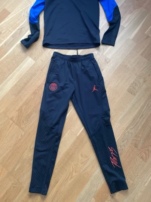 PSG x Jordan träningsset svart/blå - Snyggt träningsset från PSG x Jordan i svart med blå detaljer på ärmarna. Setet har röda loggor och tryck, dragkedja vid halsen och smal passform. Materialet är mjukt och stretchigt, perfekt för fotboll eller träning.